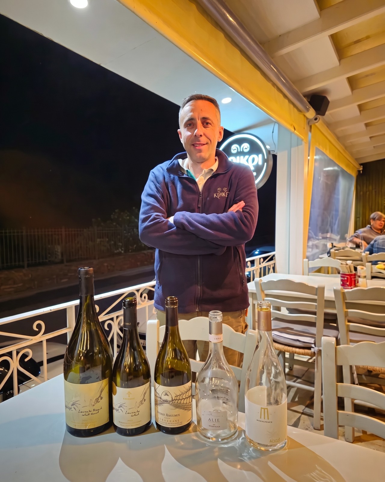 Kostas at Kri Kri in the evening, the taverna's lit sign behind him