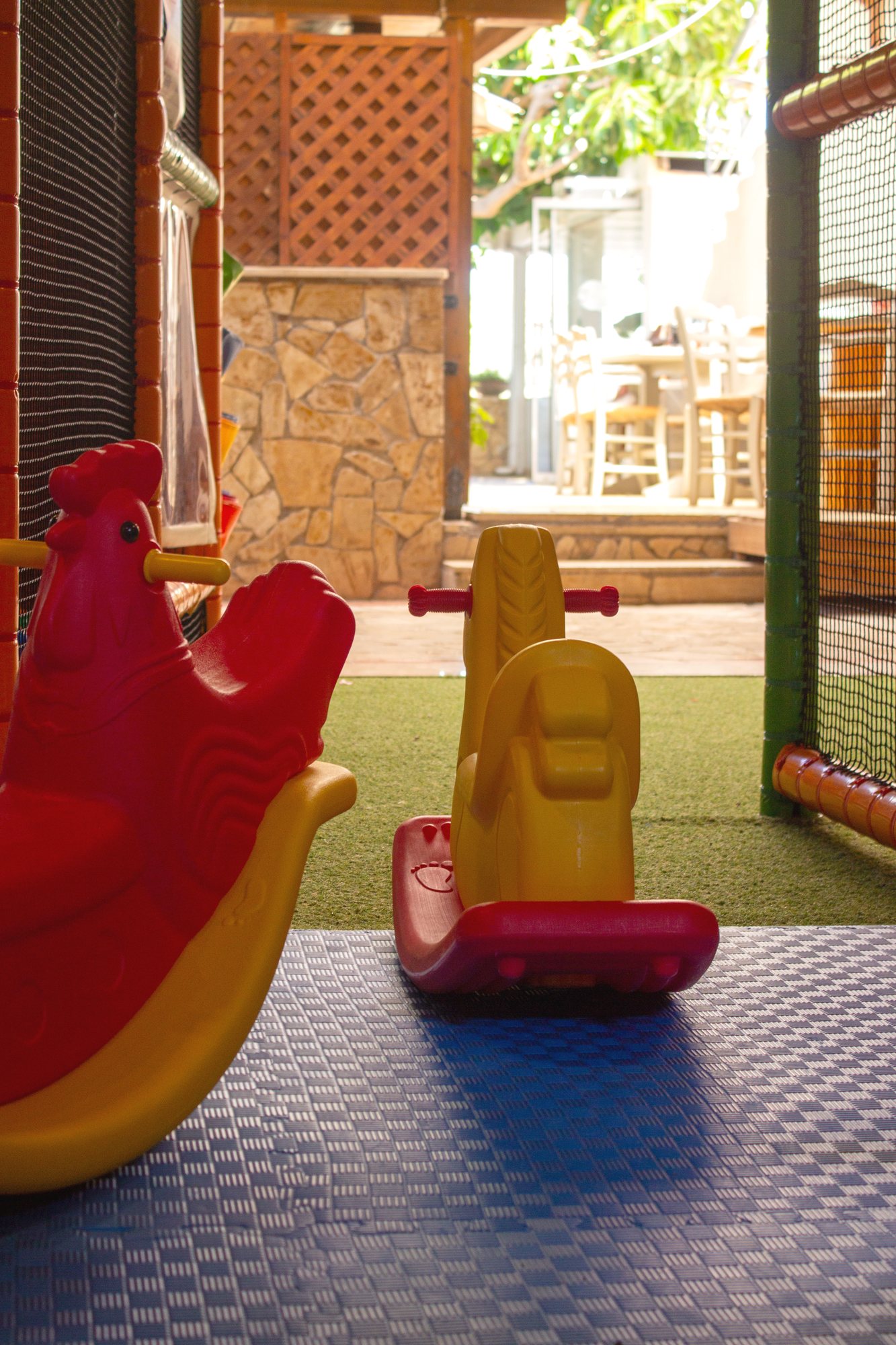 The children's playroom at Kri Kri, colourful ride-on toys on the play mat with the taverna's stone wall and tables beyond